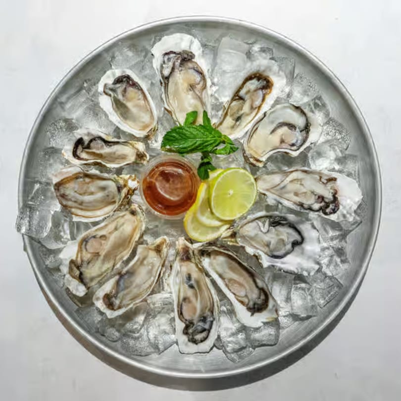 A dozen fresh oysters on a bed of ice, garnished with lime and mint, with cocktail sauce.