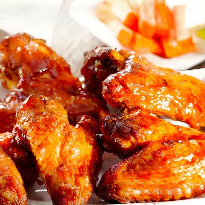 Close-up of several glossy, golden-brown chicken wings with a side of fruit in the background.