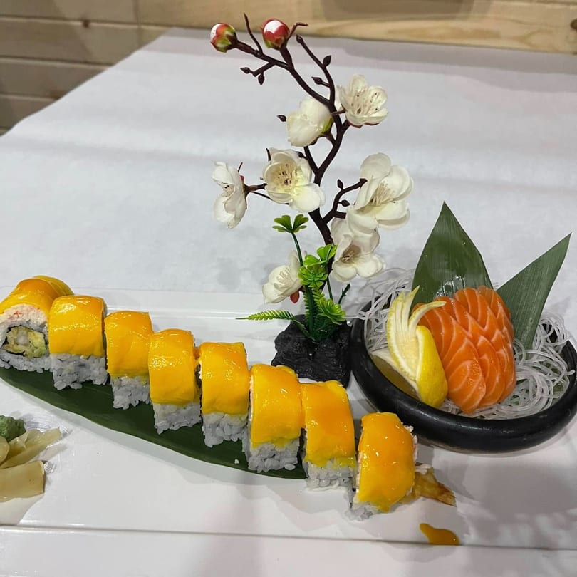 Mango sushi rolls and salmon sashimi with lemon and a decorative floral branch on a white table.